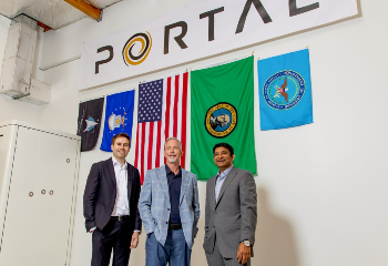 Three people stand in front of a various flags including the U.S. and Washington state flags. The logo for Portal is on the top of the wall.